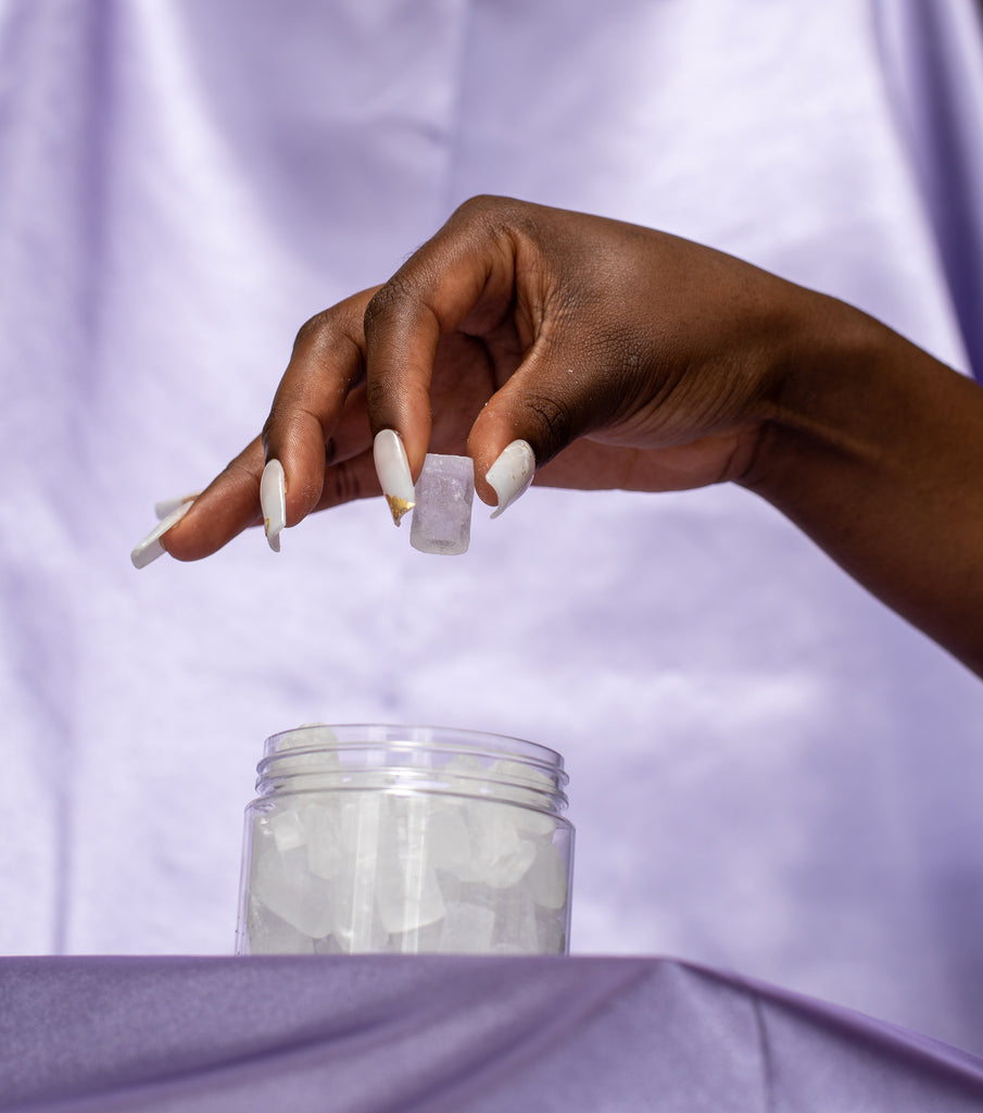 Lavender Satin backdrop. African american female hand picks up one yoni melt, a white edible crystal. Clear container of yoni melts are underneath the hand.