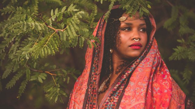Black woman with curly hair in front of a tree. She is wearing a gold head jewelry and multi colored red shawl over her head and shoulders. 
